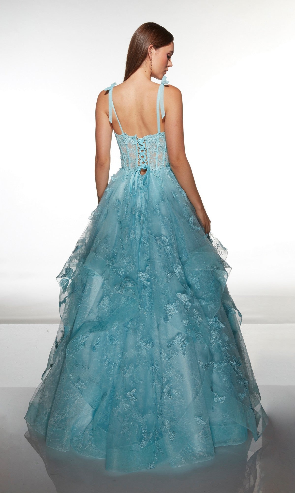 A woman stands with her back to the camera, wearing the Alyce 62199 long prom dress—a light blue gown with a corset bodice, floor-length tulle ballgown skirt, and delicate floral details—against a plain background.